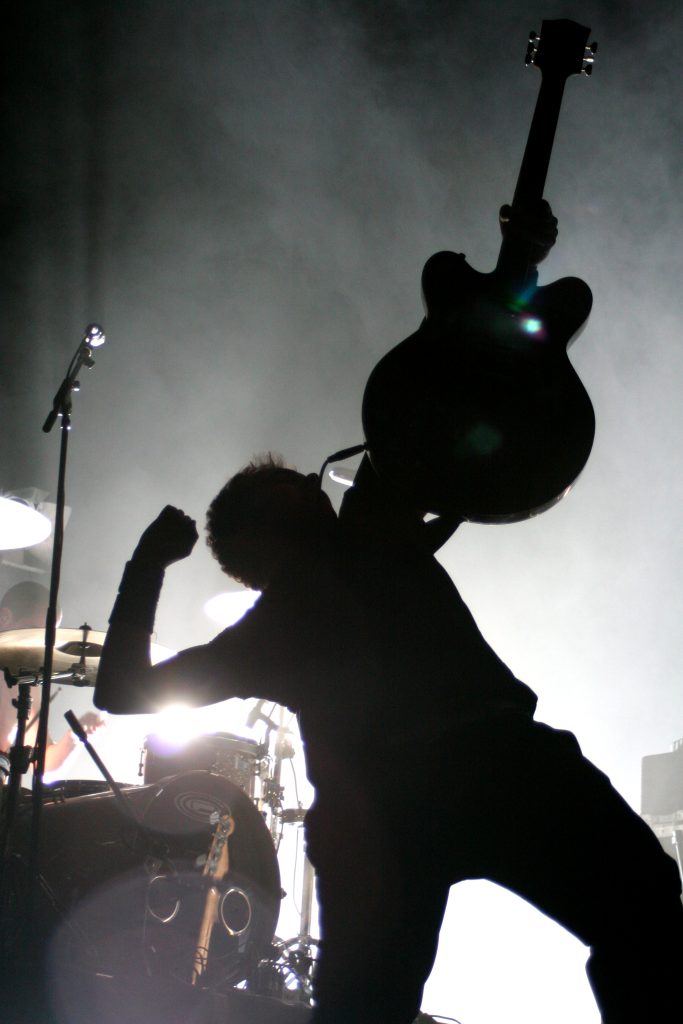 Silhouette en contre-jour d'un musicien du groupe Dionysos brandissant sa guitare à bout de bras, photographie de concert graphique et rock à Toulouse par Well Com Studio Graphique à Albi.