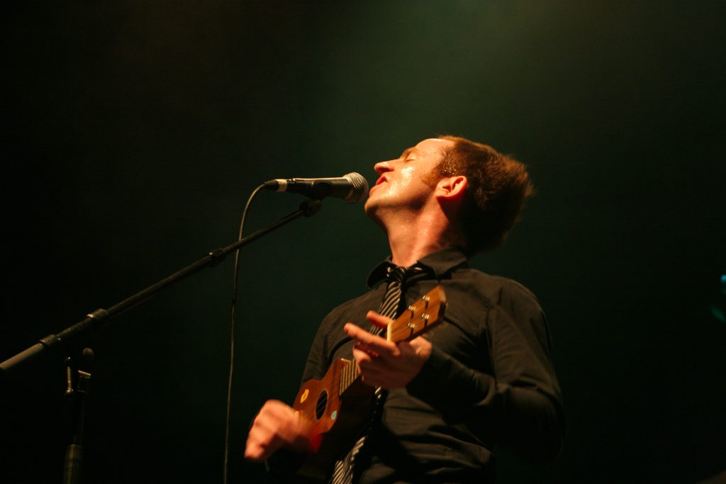 Portrait gros plan de Mathias Malzieu (groupe Dionysos) chantant les yeux fermés avec son ukulélé, éclairage dramatique sur scène à Toulouse (2013), par Well Com Studio Graphique à Albi.