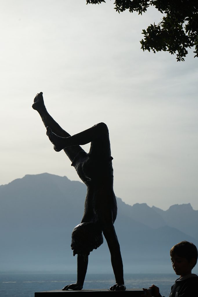 Silhouette de statue d'enfant faisant le poirier en contre-jour devant le Lac Léman et les montagnes, photographie artistique et jeu d'ombres par Well Com Studio Graphique à Albi.