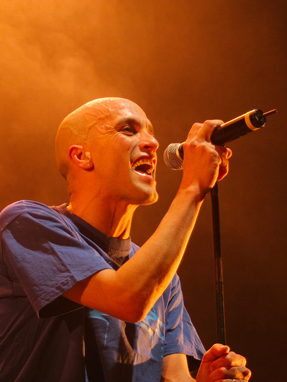 Photographie de concert du chanteur Magyd Cherfi (groupe Zebda) sur scène à Toulouse lors d'un événement organisé par l'AFEV. Portrait scénique dynamique avec éclairage chaud par Well Com Studio Graphique à Albi.