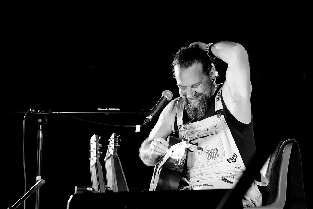 Photographie de concert noir et blanc du musicien Will Barber sur scène (souriant avec sa guitare lap steel) lors du festival Ramène ta Chaise 2023 à Puygouzon. Reportage événementiel par Well Com Studio Graphique à Albi.