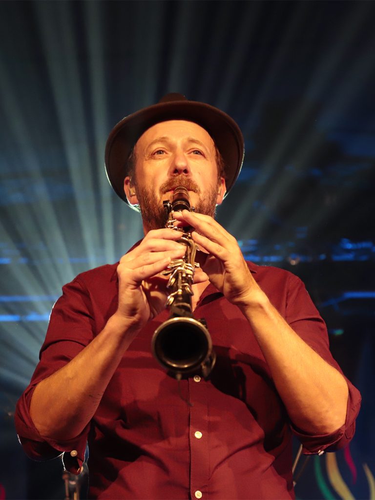 Portrait d'un musicien jouant de la clarinette (groupe Debout sur le Zinc) avec chapeau, éclairé par des faisceaux lumineux en arrière-plan. Photographie de concert par Well Com Studio Graphique à Albi.
