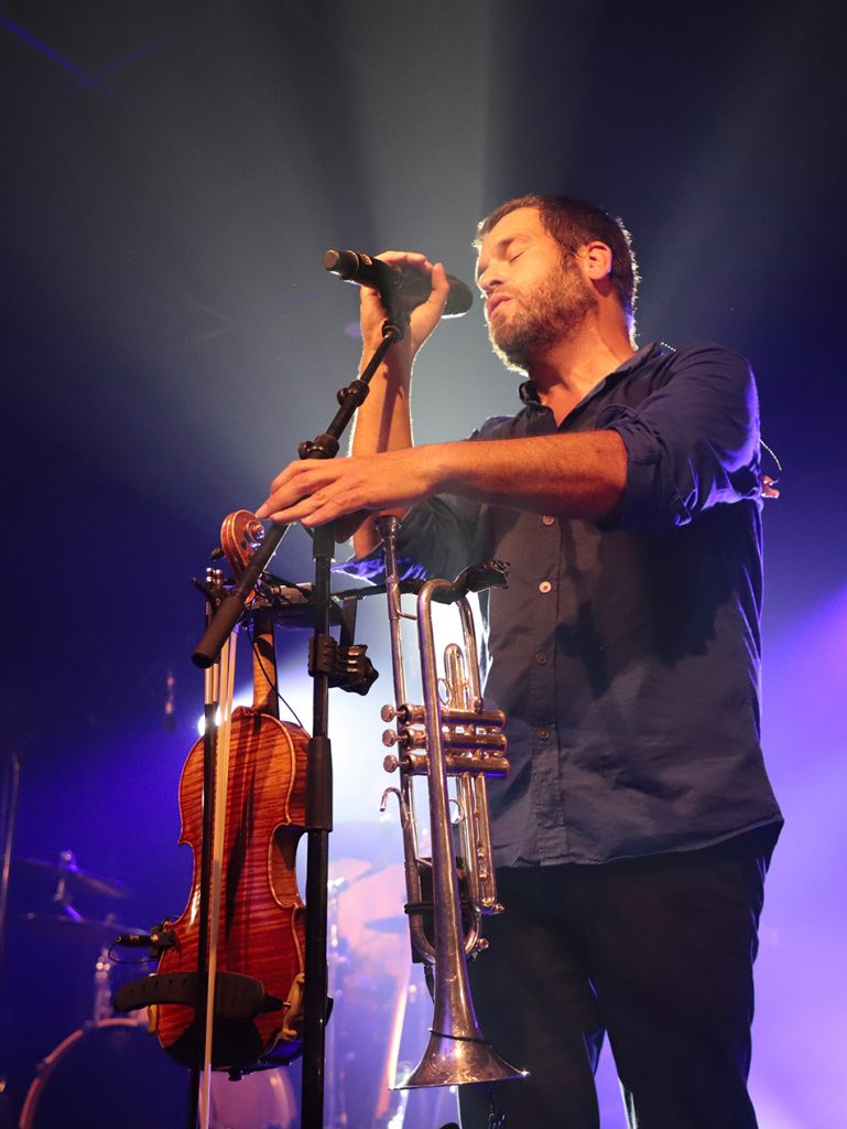 Portrait du chanteur du groupe Debout sur le Zinc chantant les yeux fermés, entouré d'un violon et d'une trompette sur son pied de micro. Photographie de concert au Festival Un Bol d'Air à Puygouzon par Well Com Studio Graphique à Albi.