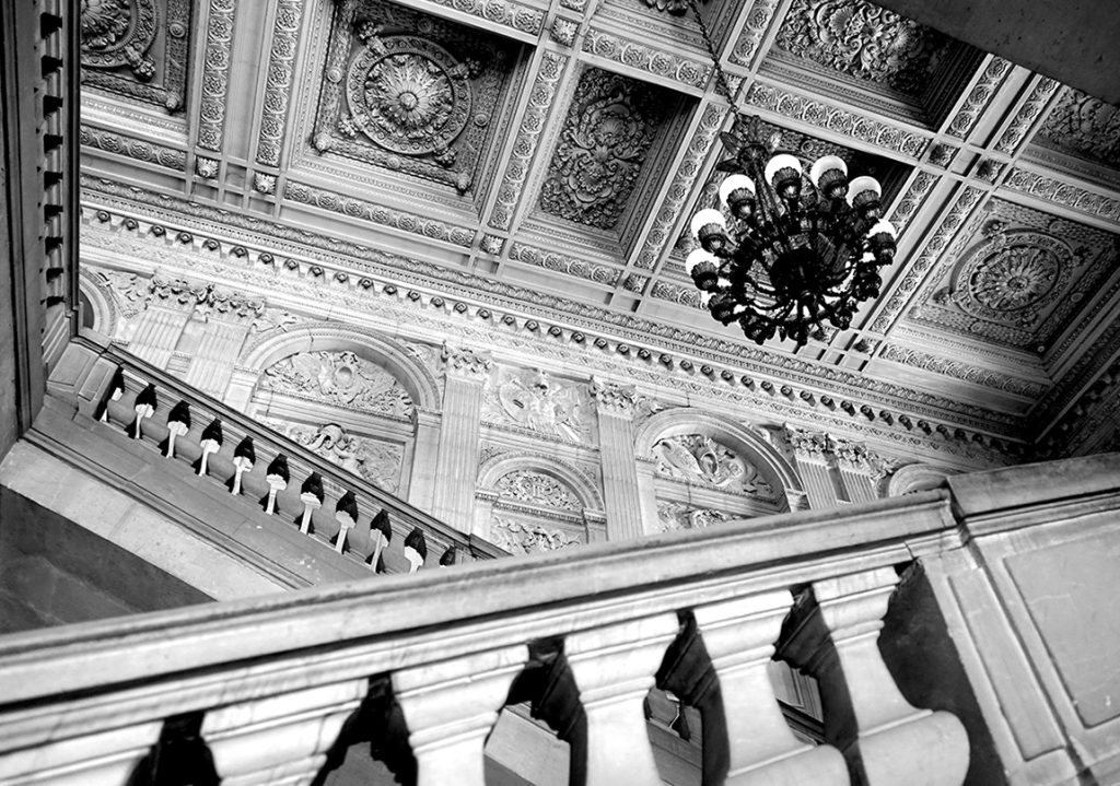 Vue en contre-plongée de l'intérieur du Musée du Louvre à Paris : plafond à caissons sculptés, lustre monumental et rampe d'escalier en pierre. Photographie d'architecture noir et blanc par Well Com Studio Graphique à Albi.