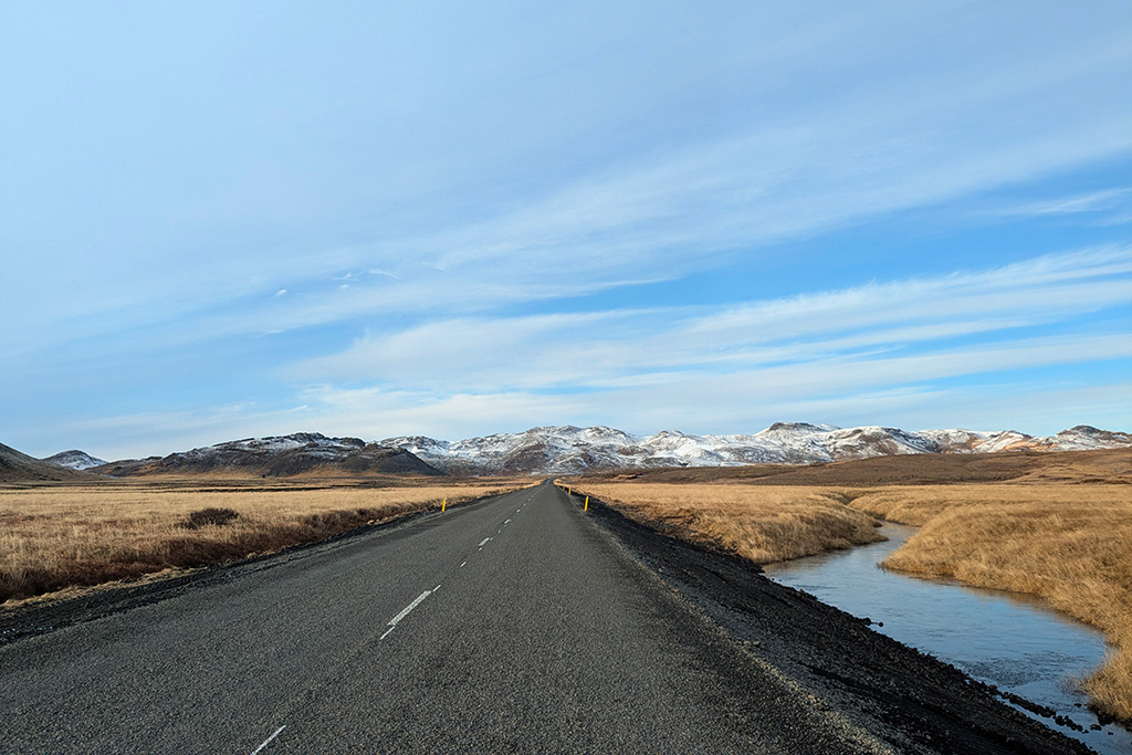 Route asphaltée vide traversant un paysage islandais aride sous un ciel bleu nuageux, avec des montagnes enneigées au loin et un cours d'eau sur la droite, photographie de road trip par Well Com Studio Graphique à Albi.