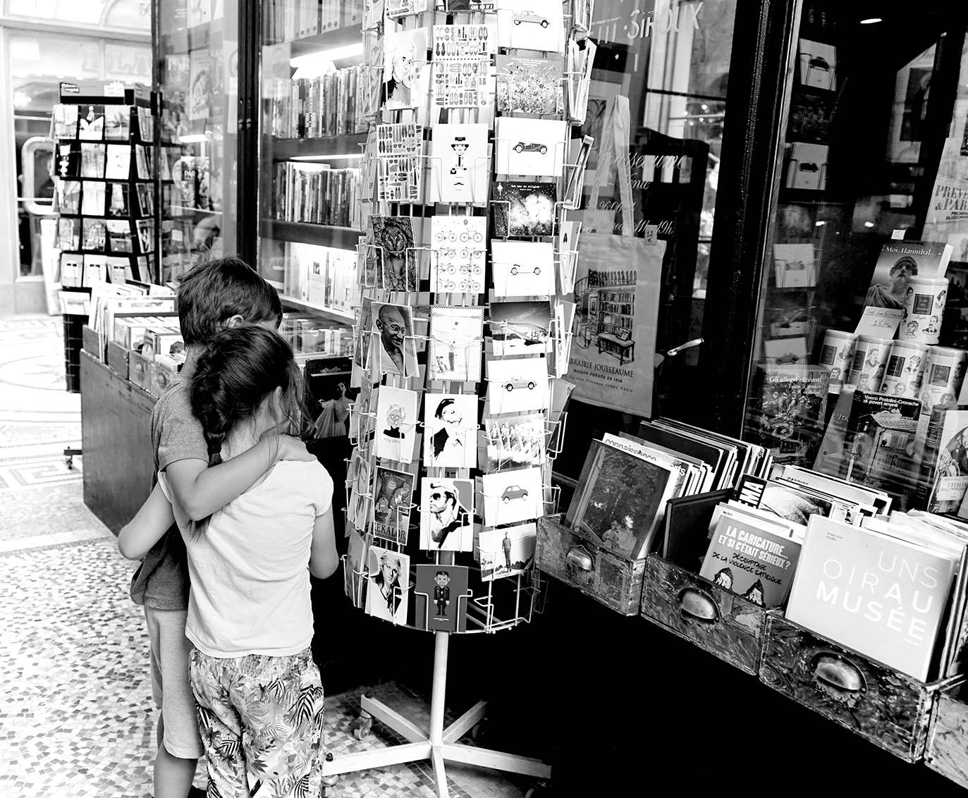 Photographie noir et blanc de deux enfants regardant un présentoir de cartes postales devant une librairie au Passage du Grand Cerf à Paris, scène de vie urbaine par Well Com Studio Graphique à Albi.