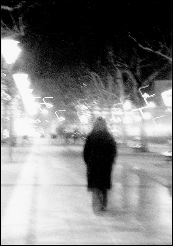 Photographie argentique noir et blanc de nuit à Albi : silhouette floue marchant vers la Place du Vigan entourée de traînées de lumière (effets de mouvement), par Well Com Studio Graphique à Albi.