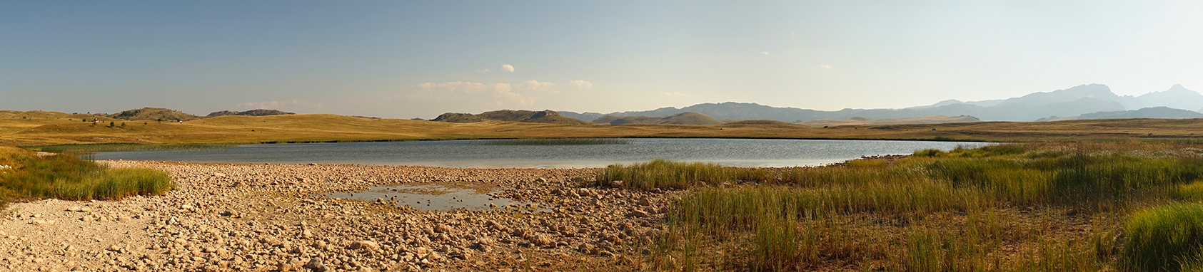Panorama du plateau des lacs au Monténégro : contraste entre les herbes dorées, l'eau bleue et les montagnes en arrière-plan. Photographie de paysage et inspiration couleur par Well Com Studio Graphique à Albi.