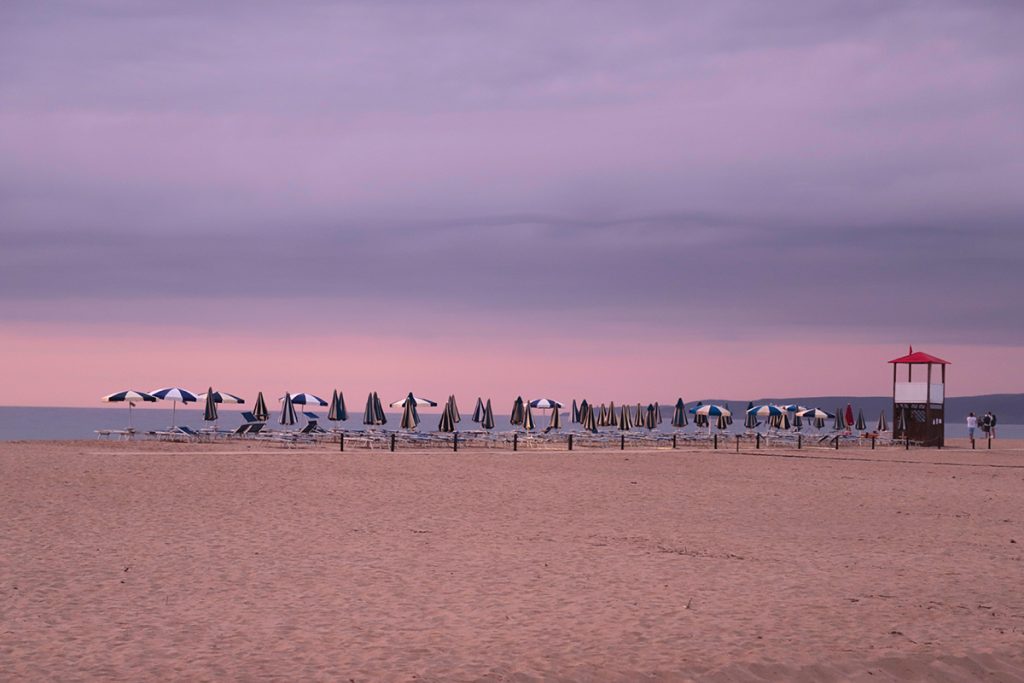 Plage de Piscinas en Sardaigne au crépuscule : rangées de parasols fermés sous un ciel dégradé rose et violet, photographie d'ambiance et inspiration couleur par Well Com Studio Graphique à Albi.